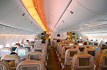 客機機艙。兩排座位排列在兩條走道之間。每個座椅靠背都有一個個人顯示器，其光線從側壁和頭頂行李箱照射進來。