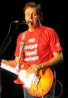 McCartney, in his late sixties, playing an orange electric guitar and wearing a red shirt that bears, in white writing, the words "no more land mines". His eyes are closed.