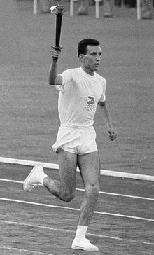Athlete in white shorts and T-shirt running on an athletics track with the Olympic torch aloft in his right hand.