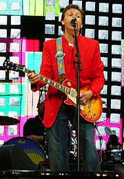 A colour photograph of McCartney, wearing a red coat and blue jeans playing an electric guitar and singing, while performing live on a stage.