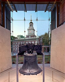 The Liberty Bell hangs in a glass-backed structure, with a brick, 18th-century building with a steeple visible in the background.