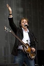 Colour photograph of McCartney, in his sixties, playing a Höfner 500/1 electric bass. He wears a black buttoned-up suit jacket with black pants and white shirt.
